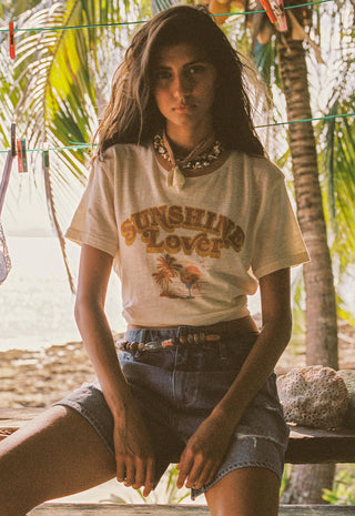 A woman sitting outdoors on a wooden bench wearing a cream graphic t-shirt with 'SUNSHINE LOVER' printed on the front