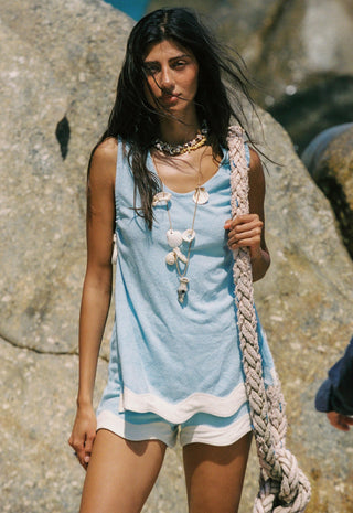 A woman standing outdoors against large rocks, wearing a light blue sleeveless top with white shell-like decorative details, white shorts, layered necklaces, and holding a thick braided rope.