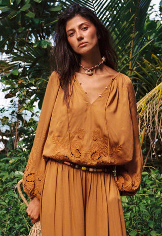 a-woman-wearing-a-bohemian-style-mustard-yellow-embroidered-top-and-matching-pleated-skirt-standing-outdoors-in-front-of-lush-green-tropical-foliage