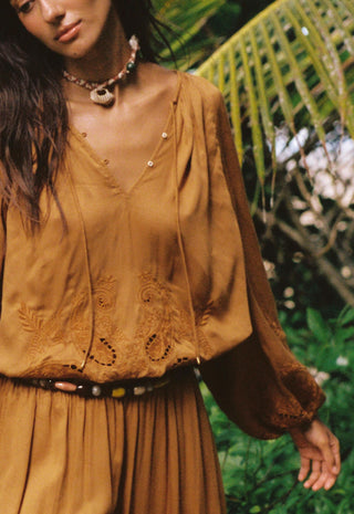 a-woman-wearing-a-loose-earthy-brown-bohemian-style-blouse-with-embroidered-details-and-a-matching-pleated-skirt-styled-with-a-patterned-necklace-and-belt-set-against-a-lush-green-tropical-background