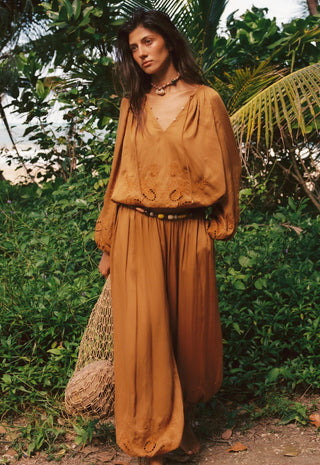 a-woman-stands-outdoors-in-a-lush-green-setting-wearing-a-matching-rust-brown-bohemian-long-sleeve-top-and-wide-leg-skirt-set-with-a-black-belt-she-is-holding-a-woven-straw-bag