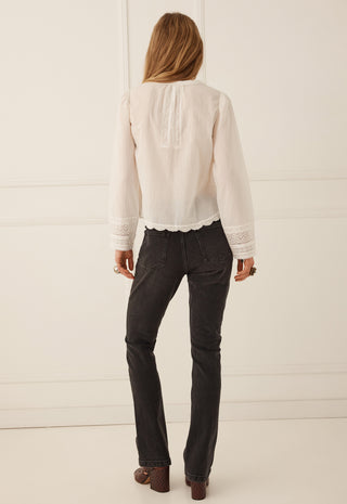 A woman viewed from behind wearing a cream-colored long-sleeve blouse with lace trim and dark charcoal gray jeans, standing against a white paneled wall.