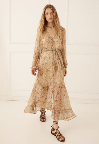 A woman modeling a long-sleeved, V-neck maxi dress with a beige and brown floral pattern, paired with brown lace-up sandals, set against a white paneled wall.
