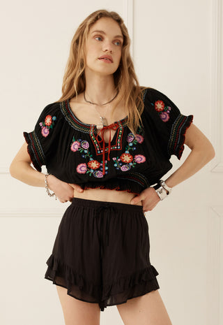a-woman-modeling-a-black-short-sleeve-crop-top-with-colorful-floral-embroidery-and-a-red-tie-detail-paired-with-black-ruffled-shorts-against-a-white-paneled-wall