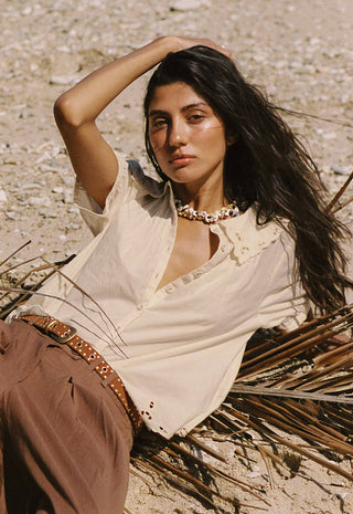 a-woman-reclining-outdoors-on-a-sandy-ground-wearing-a-cream-linen-shirt-brown-pants-and-a-studded-brown-belt-accessorized-with-a-chunky-gold-necklace