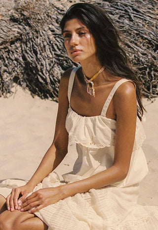 a-woman-sitting-on-a-sandy-beach-wearing-a-cream-colored-ruffled-spaghetti-strap-dress-and-a-gold-necklace