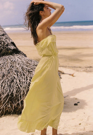 a-woman-wearing-a-strapless-pale-yellow-maxi-dress-stands-on-a-sandy-beach-with-the-ocean-and-a-thatched-palm-tree-in-the-background