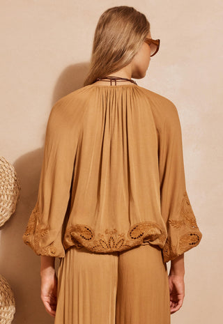 a-woman-shown-from-the-back-wearing-a-camel-brown-pleated-blouse-with-embroidered-lace-sleeves-and-matching-pleated-trousers-set-against-a-warm-beige-textured-wall