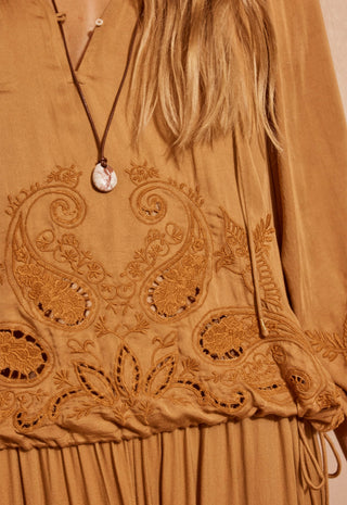 close-up-of-a-tan-embroidered-dress-with-paisley-and-floral-cutout-embroidery-featuring-a-brown-cord-necklace-with-a-round-shell-pendant-worn-by-a-model