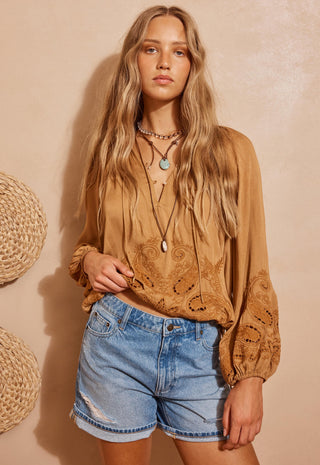 a-woman-modeling-a-tan-bohemian-long-sleeve-blouse-with-intricate-floral-embroidery-and-eyelet-cutouts-paired-with-light-wash-denim-shorts-set-against-a-textured-beige-wall-with-woven-baskets