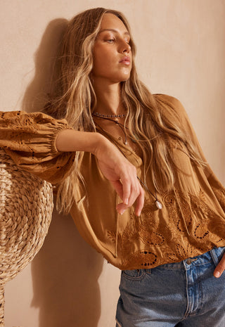 a-woman-wearing-a-tan-eyelet-lace-blouse-with-puff-sleeves-and-blue-jeans-leaning-against-a-textured-beige-wall-next-to-a-woven-straw-hat