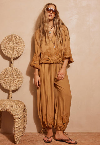a-woman-models-a-matching-tan-embroidered-two-piece-set-consisting-of-a-loose-long-sleeve-top-and-high-waisted-harem-pants-styled-with-sunglasses-layered-necklaces-and-flip-flops-against-a-textured-beige-wall-next-to-a-woven-chair