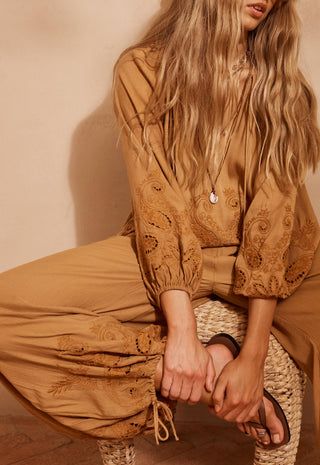 a-woman-wearing-a-matching-tan-embroidered-blouse-and-wide-leg-pants-seated-on-a-woven-stool-against-a-warm-beige-wall