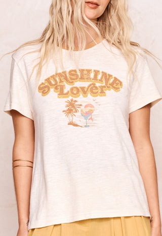 A woman wearing a light cream heathered t-shirt with a retro-style graphic that reads 'SUNSHINE LOVER' in gold/yellow letters above a small beach illustration. The background is a warm, textured off-white wall.