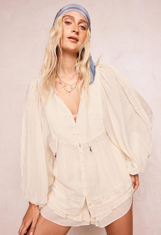 a-woman-modeling-a-cream-colored-long-sleeved-bohemian-style-romper-with-pleated-details-and-lace-trim-paired-with-a-light-blue-patterned-headband-against-a-neutral-beige-background