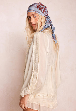 a-woman-wearing-a-cream-colored-semi-sheer-long-sleeve-blouse-with-lace-trim-and-a-blue-paisley-bandana-headscarf-photographed-against-a-textured-beige-background