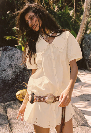 a-woman-outdoors-wearing-a-cream-colored-short-sleeve-button-front-dress-with-eyelet-trim-and-a-matching-belt-standing-among-rocks-and-tropical-plants
