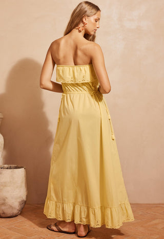 a-woman-wearing-a-strapless-yellow-maxi-dress-with-ruffled-trim-and-a-tie-waist-shown-from-the-back-in-a-warm-toned-indoor-setting