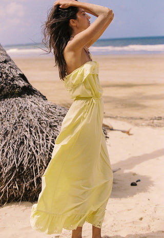 A woman stands on a sandy beach wearing a strapless pale yellow maxi dress with a gathered waist and ruffled hem. A large thatched palm tree and ocean are visible in the background.