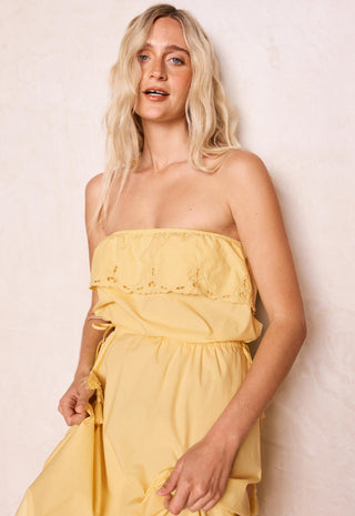 A blonde woman wearing a yellow strapless midi dress with a ruffled eyelet bodice, standing against a textured off-white wall.