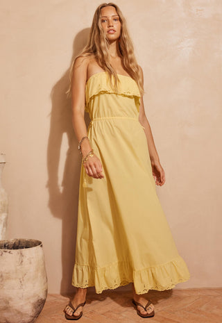 A woman modeling a strapless yellow maxi dress with ruffle details, standing against a textured beige wall in a warm, rustic setting.