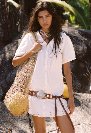 A woman stands outdoors on rocky ground wearing a white short-sleeve button-front shirt dress with eyelet trim, a brown woven belt with a large silver buckle, layered necklaces, and a large beige mesh tote bag containing yellow coconuts.