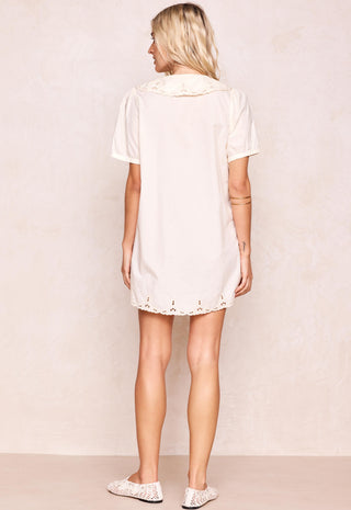A woman shown from the back wearing a short-sleeve cream-colored tunic dress with lace trim at the collar and hem, paired with white lace-up flats, set against a textured beige studio backdrop.