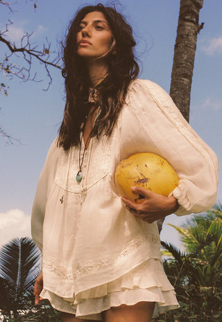 a-woman-outdoors-wearing-a-loose-white-bohemian-style-blouse-and-holding-a-yellow-coconut-under-her-arm-with-palm-trees-and-blue-sky-in-the-background
