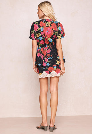 a-woman-viewed-from-the-back-wearing-a-short-sleeve-black-dress-with-a-vibrant-multicolor-floral-print-and-a-white-lace-hem-paired-with-gray-block-heel-sandals-against-a-textured-beige-wall