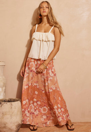 a-woman-modeling-a-white-ruffled-camisole-top-paired-with-a-long-peach-pink-floral-maxi-skirt-standing-against-a-warm-beige-textured-wall