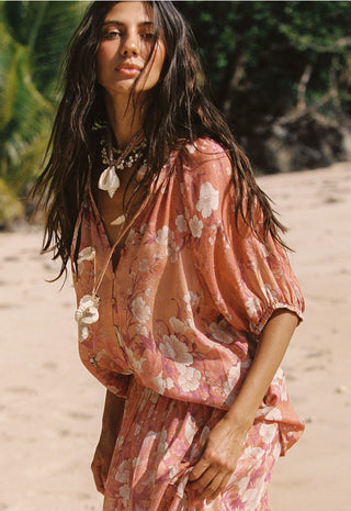 a-woman-on-a-sandy-beach-wearing-a-loose-dusty-pink-floral-dress-with-voluminous-short-sleeves-and-a-draped-neckline