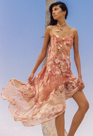 a-woman-wearing-a-flowing-floral-slip-dress-in-soft-pink-and-peach-tones-posed-outdoors-against-a-clear-blue-sky-with-a-palm-tree-visible-behind-her