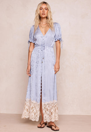 A woman modeling a light blue maxi dress with short puff sleeves, a V-neckline, and a lace hem, standing against a textured beige wall.