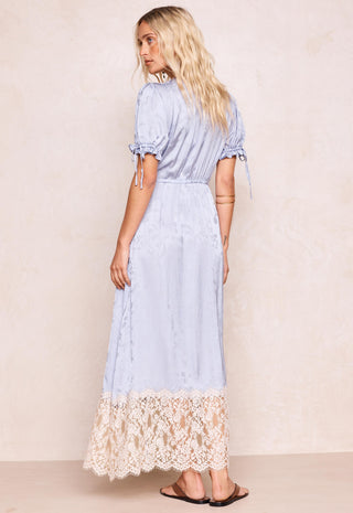 A woman modeling a light blue maxi dress with short puff sleeves, side tie details, and a cream lace hem, shown from the back against a textured beige wall.