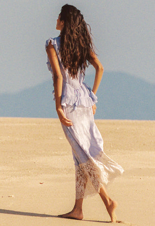 a-woman-with-long-dark-hair-stands-barefoot-on-a-sandy-beach-viewed-from-behind-wearing-a-flowing-light-blue-and-white-dress-with-lace-trim