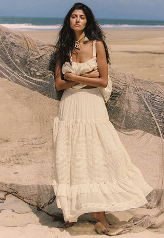 a-woman-stands-on-a-sandy-beach-wearing-a-long-cream-maxi-dress-with-a-tiered-ruffle-skirt-a-large-beige-fishing-net-is-draped-across-the-beach-behind-her-with-the-ocean-and-blue-sky-in-the-background