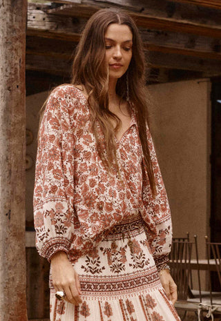 a-woman-wearing-a-bohemian-style-long-sleeve-maxi-dress-with-a-rust-brown-and-cream-floral-pattern-standing-in-front-of-a-rustic-wooden-structure