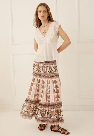 a-woman-modeling-a-bohemian-style-two-piece-outfit-featuring-a-white-sleeveless-lace-trimmed-top-and-a-long-cream-maxi-skirt-with-brown-floral-and-geometric-patterns-paired-with-brown-strappy-sandals-against-a-white-paneled-wall