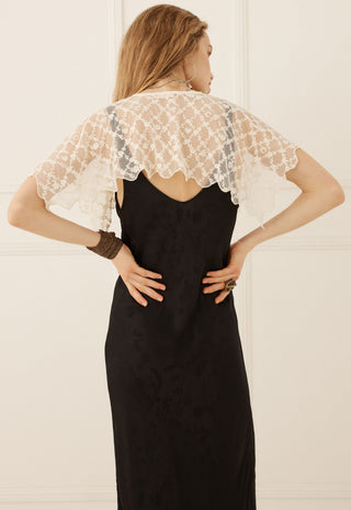 a-woman-viewed-from-the-back-wearing-a-black-slip-dress-with-a-sheer-white-lace-flutter-cape-overlay-set-against-a-light-cream-paneled-wall